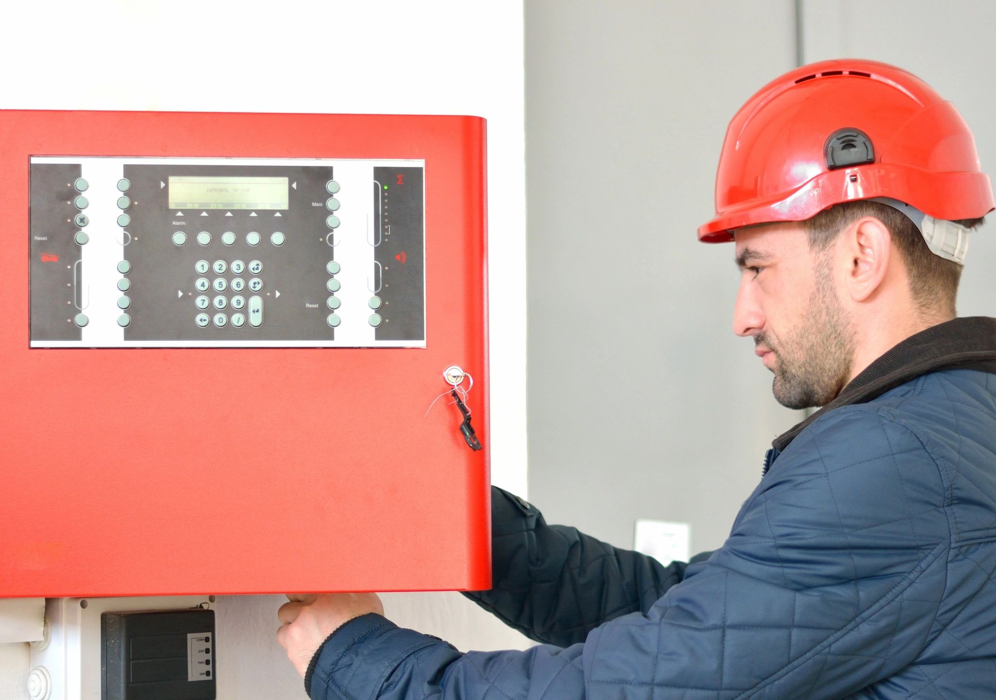 A man in red hard hat holding on to a fire alarm.